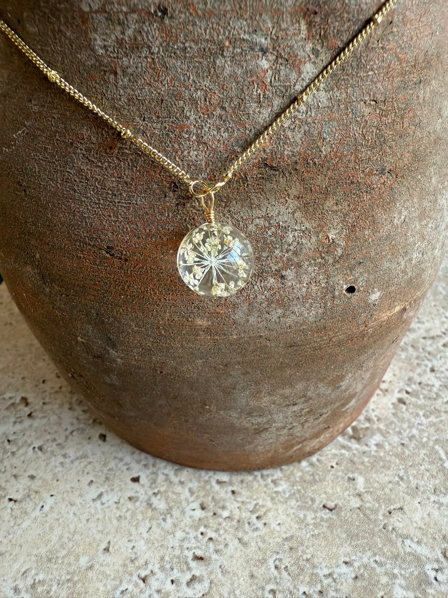 Collier pendentif rond en résine avec fleur séchée blanche encapsulée, chaîne fine dorée, bijou artisanal élégant posé devant vase en terre cuite rustique.